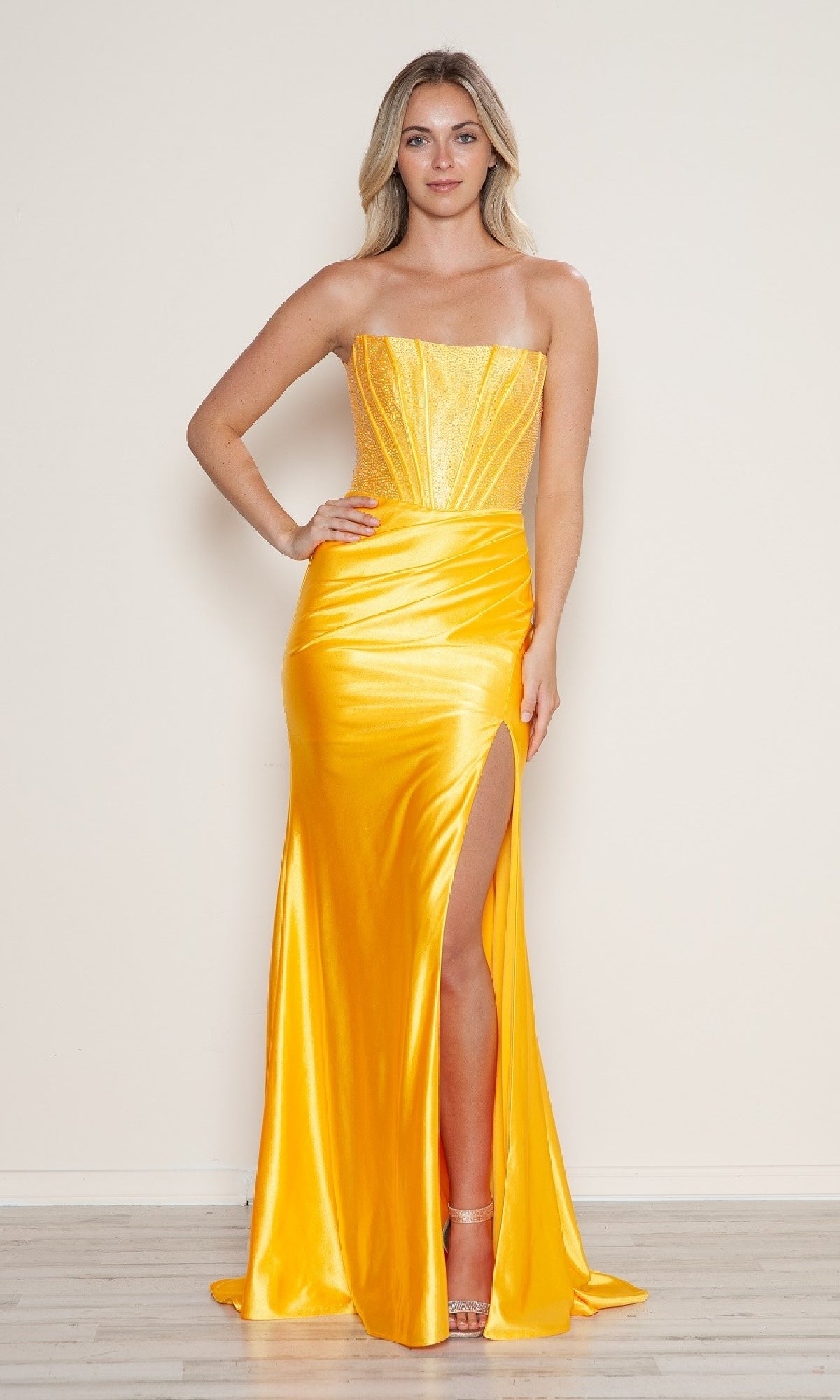 Woman wearing the Off-Shoulder Long Corset Prom Dress: Poly USA 9656, a bright yellow satin gown with a fitted bodice and high slit, standing against a plain light-colored wall.
